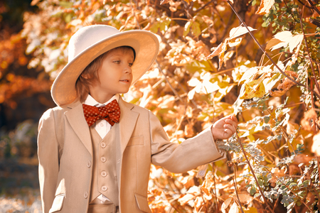 Portrait of a cute funny little boy in elegant white suit and white hat walking through a beautiful autumn park. Retro style. Children's fashion.の写真素材