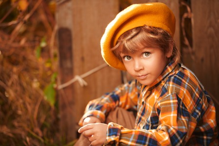 Autumn season. Cute little boy sits by a wooden fence in a beautiful autumn park. Retro style. Children's fashion.の写真素材
