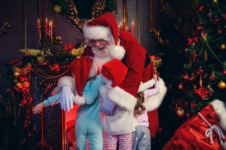 Santa Claus cuddles with little girls at home. Merry Christmas and Happy New Year. Miracle time.の写真素材
