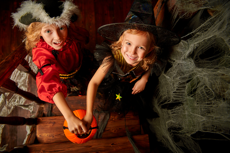 Two happy girls in carnival costumes in the old wooden house with pumpkin basket. Fairy tales on Halloween. Trick or treat.の写真素材