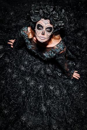 Scary and beautiful Calavera Catrina looking up at camera over dark background. Fashion model with sugar skull makeup. Dia de los muertos. Day of The Dead. Halloween.の写真素材