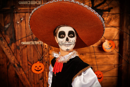 Portrait of a funny teen boy with sugar skull makeup looking at the camera in surprise. Wooden Halloween decoration. Dia de los muertos. Day of the dead.の写真素材