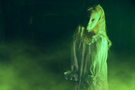 Lonely little girl in a white nightgown stands in mysterious green light. Black background....