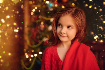 Portrait of a happy little girl against the background of a beautiful Christmas tree in a wooden house. The atmosphere of magic, lights and sparkle is around.の写真素材