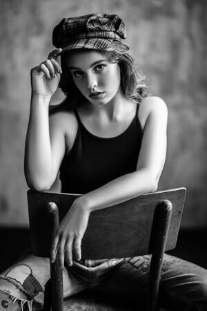A portrait of a young beautiful woman in a cap sitting on the chair in the studio. Casual fashion, beauty.の写真素材