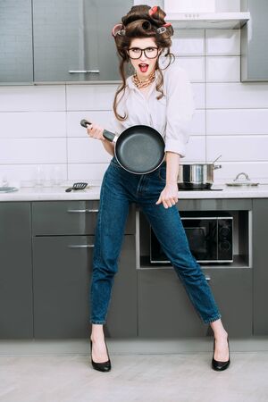 An attractive girl in everyday clothes and curlers stands in the kitchen with a frying pan. Fashion home shot. Full length portrait.の写真素材