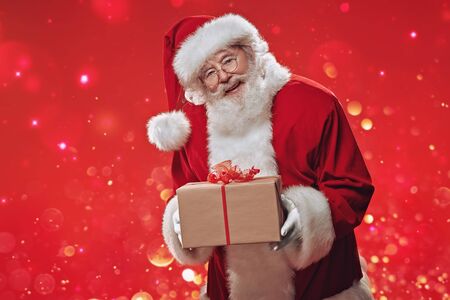 Jolly Santa Claus holds a gift box  over festive red background. The atmosphere of celebration and magic, lights and sparkling around. Merry Christmas and Happy New Year! Copy space.の写真素材