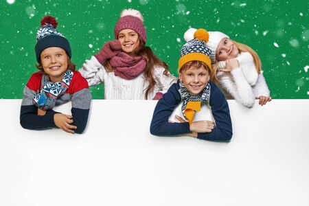 Four beautiful kids in winter clothes posing together with white board at studio. Green background. Winter fashion, winter activities. Christmas and New Year. Copy space.の写真素材