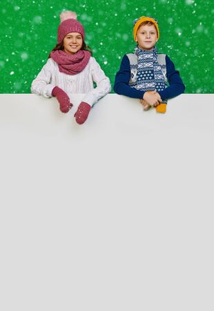 Cute boy and girl in winter clothes posing together with white board at studio. Green background. Winter fashion, winter activities. Christmas and New Year. Copy space.の写真素材