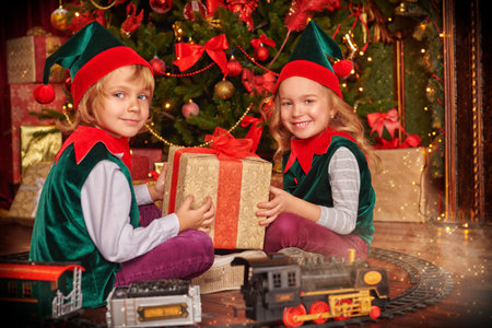 Two children sit near the Christmas tree in elf costumes and play with toys. Merry Christmas and...