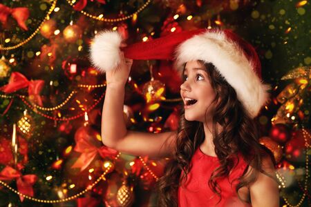 Merry Christmas and Happy New Year! Happy cute girl in red dress and Santa Claus hat celebrates Christmas by a beautiful Christmas tree.の写真素材