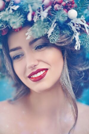 Christmas winter beauty girl. Beautiful young woman with a Christmas spruce wreath on her head, decorated with Christmas balls, berries and snowflakes smiling at camera. Snow, snowflakes.の写真素材