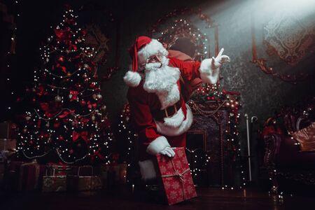 Santa Claus brought gifts for Christmas. He stands in a night room  with beautiful Christmas tree and fireplace. Christmas and New Year concept.の写真素材