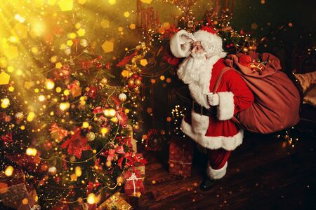 Magical Christmas. Santa Claus with a big bag of gifts stands at night in the room and looks at a beautiful Christmas tree. Lights and sparkling around. の写真素材