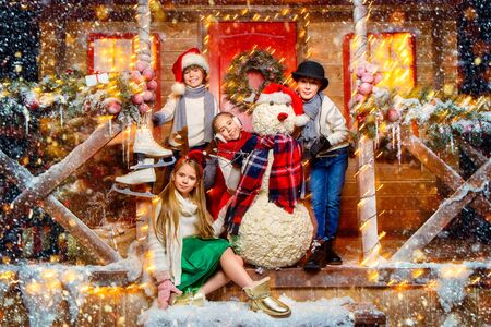 Cheerful children in winter clothes and accessories stand near the snowman on the porch of the house decorated for Christmas. Time for miracles. Merry Christmas and Happy New Year.の写真素材