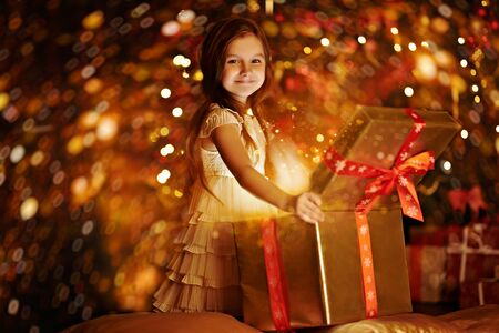 Cute little girl opens the gift box next to a beautiful Christmas tree. Christmas and New Year concept. The atmosphere of magic, lights and sparkle is around.の写真素材