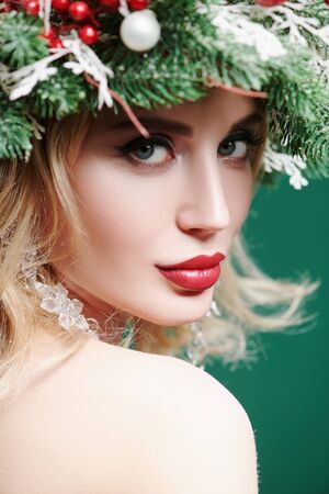Beautiful happy young woman with a Christmas pine wreath on her head, decorated with Christmas balls, berries and snowflakes. Studio portrait over xmas green background. Copy space.の写真素材