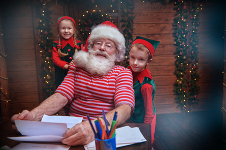 Santa Claus and little elves are sorting mail, reading letters from children. House, workshop of...