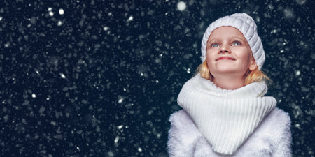 Cute girl child wearing knitted hat and a scarf stands under the snow and smiles. Kid's fashion....