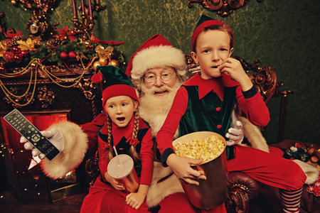 Cheerful Santa Claus and elves watching a movie with soda and popcorn in a house beautifully decorated for Christmas. Entertainment and cinema concept.の写真素材
