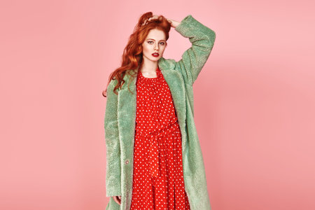 Portrait of a fashionable young woman posing in a red dress and a fur coat. Fashion shot over pink...