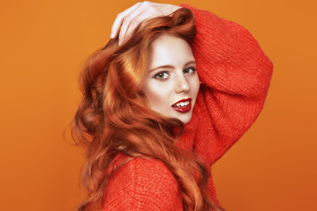 Beautiful joyful girl with bright long red hair posing at studio in red woolen sweater. Orange...