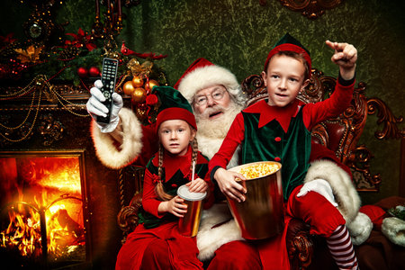 Cheerful Santa Claus and elves watching a movie with soda and popcorn in a house beautifully...