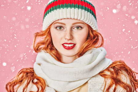Pretty red-haired girl in a knitted hat and scarf stands under snowfall and smiles. Studio portrait...