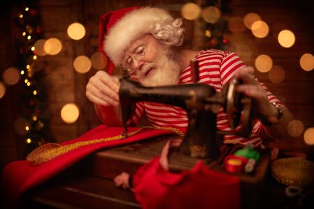 Santa Claus is preparing for Christmas at his wooden home - he sews gifts on a sewing machine. Santa Claus workshop. Christmas decoration.の写真素材