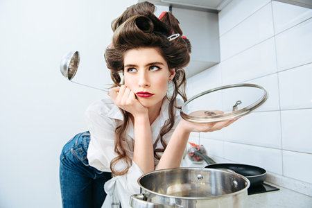 Pretty young woman with hair curlers on the head is bored standing in the kitchen with a ladle. Beauty, fashion concept. Pin-up style.の写真素材