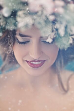 Christmas winter beauty girl. Beautiful gentle young woman with a Christmas spruce wreath on her head, decorated with Christmas balls, berries and snowflakes smiling at camera. Snow, snowflakes.の写真素材