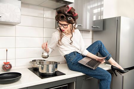 Smart pretty woman with hair curlers on her hair cooks in the kitchen and at the same time works on a laptop. Pin-up style.の写真素材