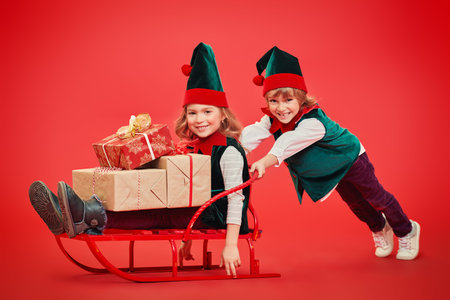 Cute little boy and girl elves helpers are pushing the sled with gifts. Studio portrait over festive...