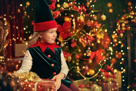Happy cute little girl sits near the Christmas tree in elf costumes and holds gift boxes. Merry Christmas and Happy New Year!の写真素材