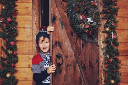 Winter activities. Scared boy child in winter clothes boy looks out from behind the door. House decorated with Christmas spruce garlands and toys. Christmas and New Year concept.の写真素材