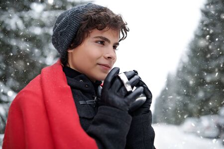 Handsome young man stands in a winter park under the snowfall and drinks hot tea. Winter season. Beauty, fashion.の写真素材