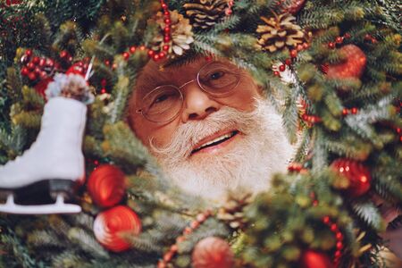 Merry Christmas and Happy New Year! Jolly Santa Claus looks through a Christmas wreath. Close-up portrait.の写真素材