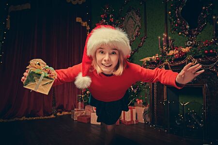 Merry Christmas and Happy New Year! Funny excited girl is jumping to the camera with a gift box. Beautiful Christmas background in the background.の写真素材