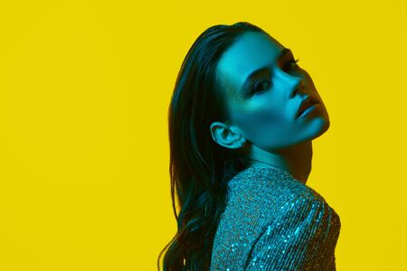 Luxurious lifestyle. Glamorous young woman with golden make-up posing in a sparkling dress in a colorful green light on a yellow background. Party style. Copy space.の写真素材
