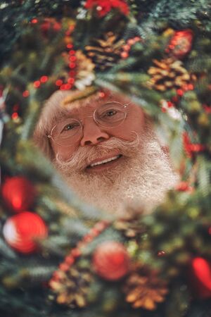 Merry Christmas and Happy New Year! Jolly Santa Claus looks through a Christmas wreath. Close-up portrait.の写真素材