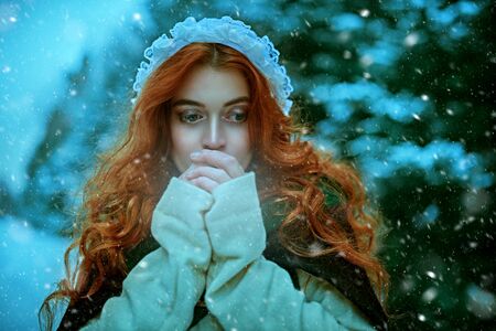 Close-up portrait of a beautiful young woman with magnificent red hair warming frozen hands in the winter forest. Middle Ages. Fairy Christmas tale. の写真素材