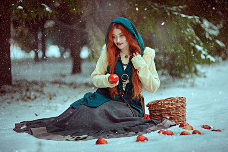 Winter fairy tale. Beautiful young woman with magnificent red hair sits on a snow in a winter forest with red apples scattered around. Historical reconstruction of the Middle Ages.の写真素材