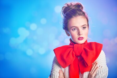 Portrait of an elegant beautiful girl in a white sweater and with a big red bow on her neck against a soft pink and blue light. Beauty, fashion concept. Winter style. Copy space.の写真素材