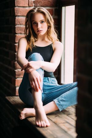 A portrait of a young beautiful woman posing on the windowsill in the room. Casual fashion, beauty.の写真素材