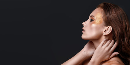 Luxurious lifestyle. Close-up portrait of a glamorous young woman with golden make-up on a grey...