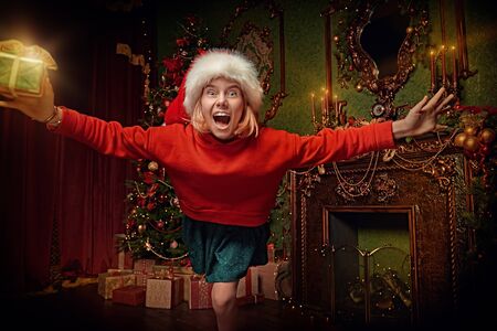 Merry Christmas and Happy New Year! Funny excited girl is jumping to the camera with a gift box. Beautiful Christmas background in the background.の写真素材