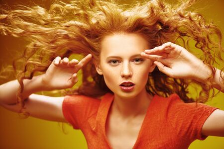 Long curly red hair. Portrait of a beautiful fashion girl with magnificent long hair in motion. Yellow background. Flying hair.の写真素材