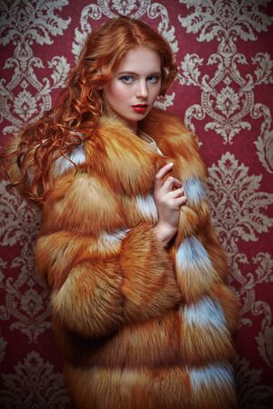 Beautiful young woman with magnificent red hair posing in a luxurious fox fur coat on a vintage background. Winter beauty fashion. Fur coat style.の写真素材