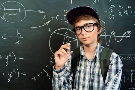 A portrait of a boy in glasses posing with a piece of chalk in front of the blackboard at school. Education, knowledge.の写真素材