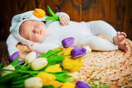 Cute newborn baby lies on a wooden background, dressed in rabbit costume next to flowers. Easter holiday. Scenery in the rustic style.の写真素材